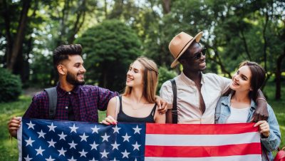 Grupo de jóvenes sonrientes sosteniendo una bandera de Estados Unidos en un parque, representando el Parole Humanitario